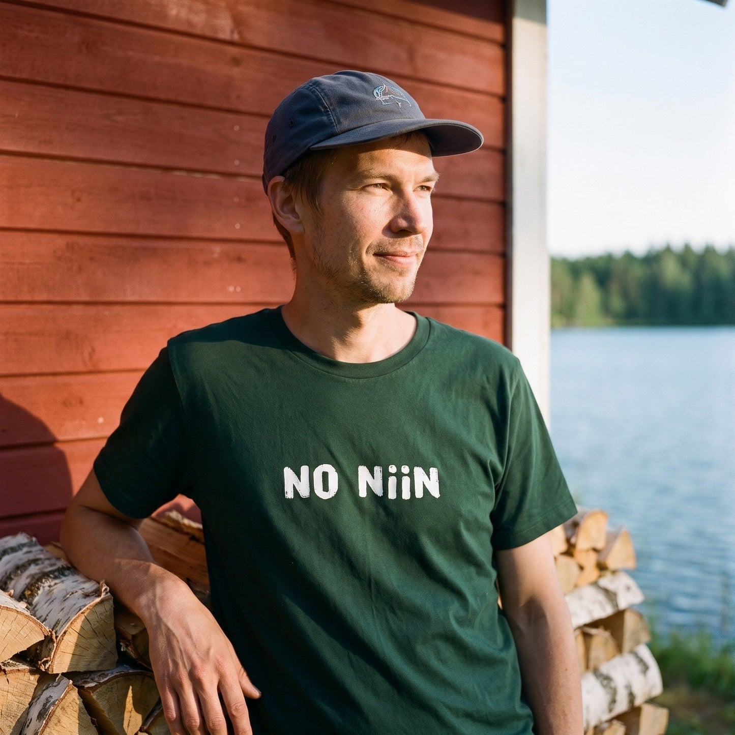 Man wearing the forest green No niin Finnish language t-shirt at a lakeside summer cottage, Very Finnish Problems