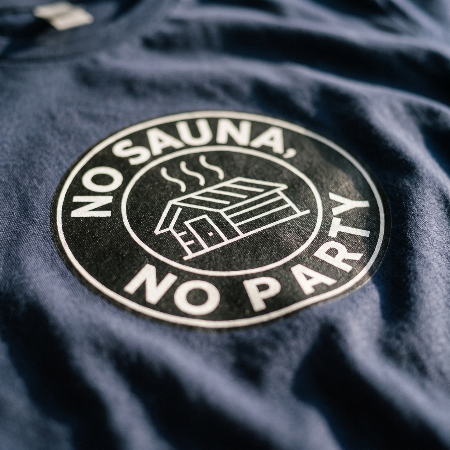 Close-up of the No sauna no party badge print on navy t-shirt fabric, showing the white lettering and sauna cabin, Very Finnish Problems