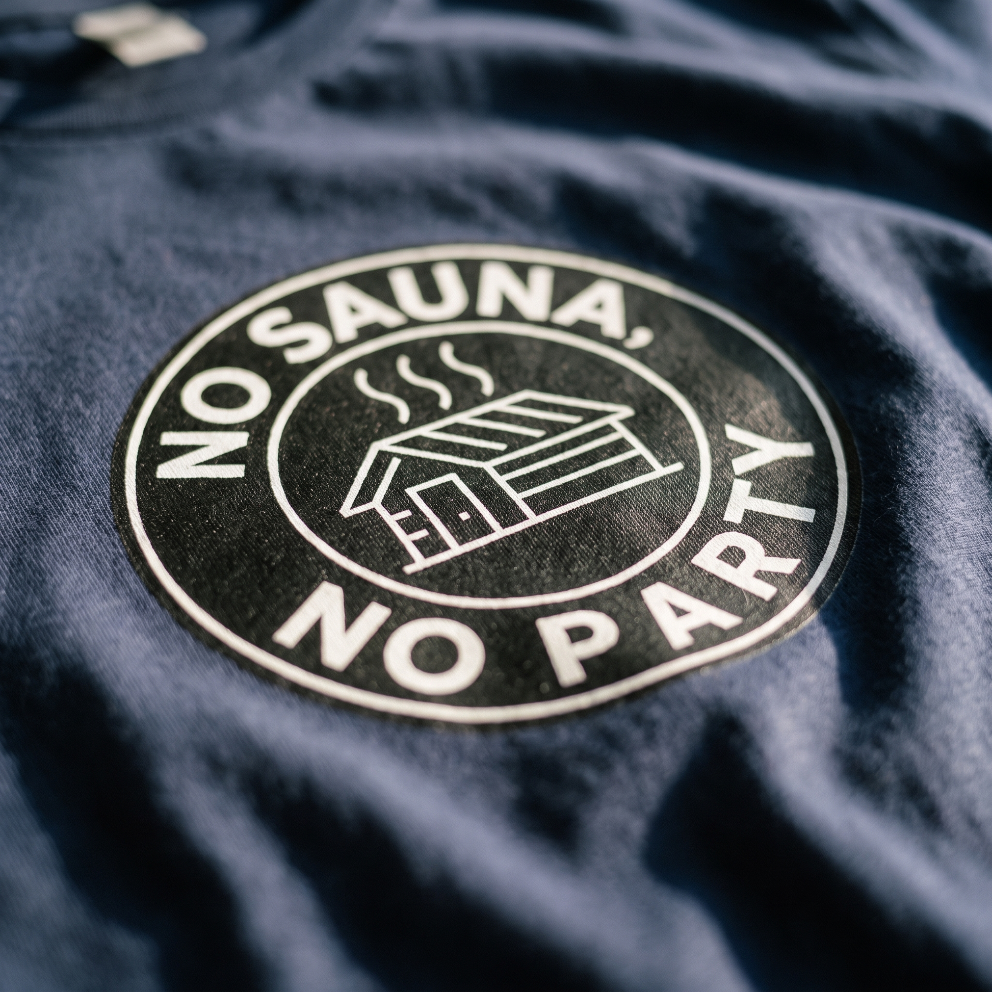 Close-up of the No sauna no party badge print on navy t-shirt fabric, showing the white lettering and sauna cabin, Very Finnish Problems