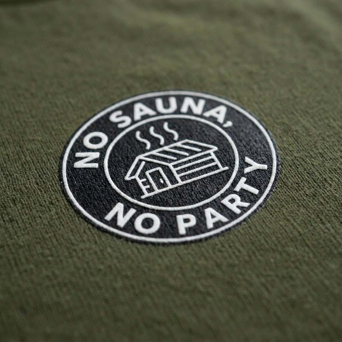 Close up of the No sauna no party badge print on military green t-shirt fabric by Very Finnish Problems