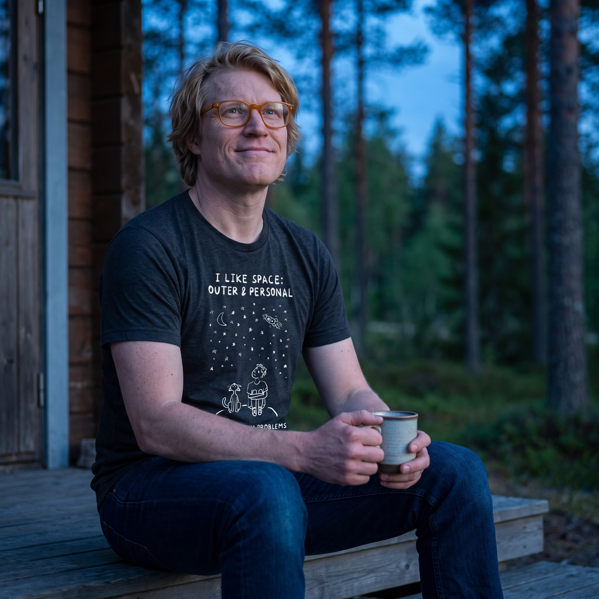 I Like Space: Outer & Personal t-shirt in dark grey, worn by Tuomas on a forest cabin doorstep at blue hour, Very Finnish Problems