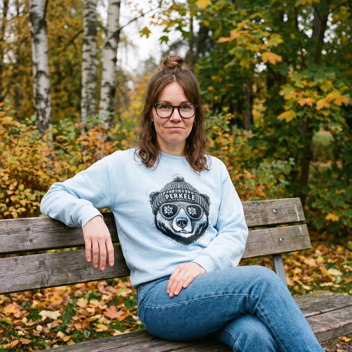Perkele Bear sweatshirt in light blue, worn by Suvi on a park bench in autumn leaves, Very Finnish Problems