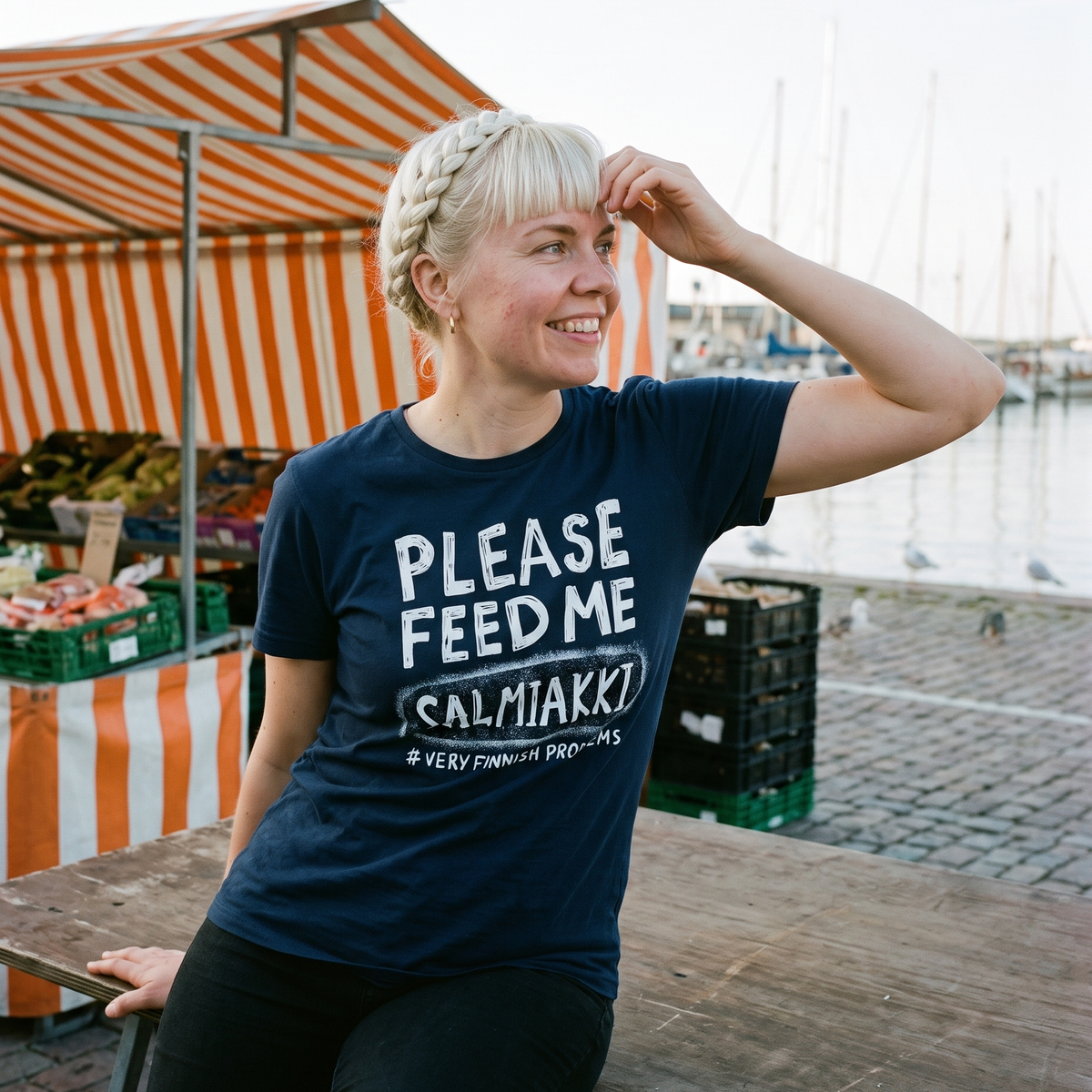 Please Feed Me Salmiakki t-shirt in navy, worn by Venla sitting on a stall table at Kauppatori market in Helsinki, Very Finnish Problems