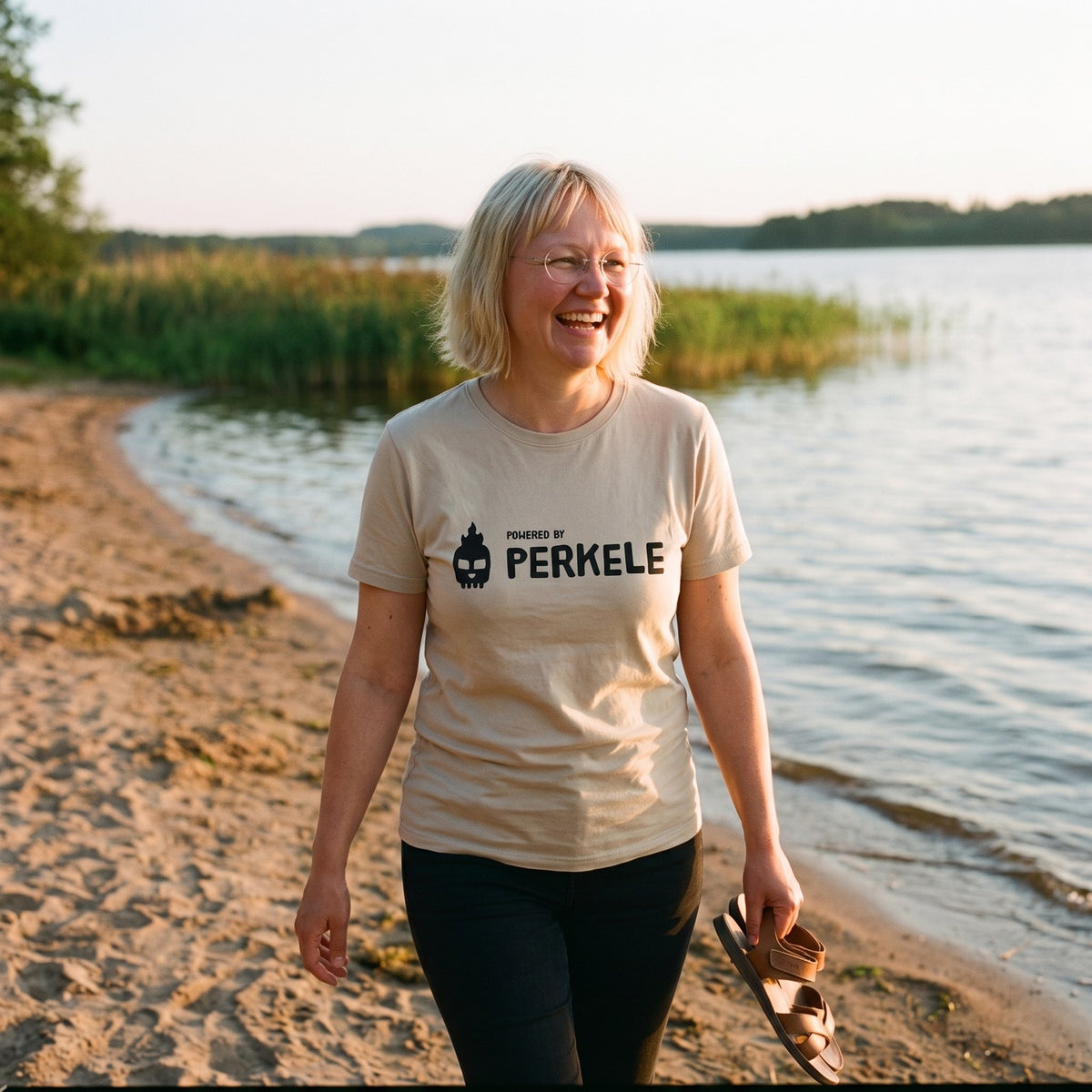 Powered by Perkele organic t-shirt in desert dust, worn by Marika on a lakeside beach, Very Finnish Problems