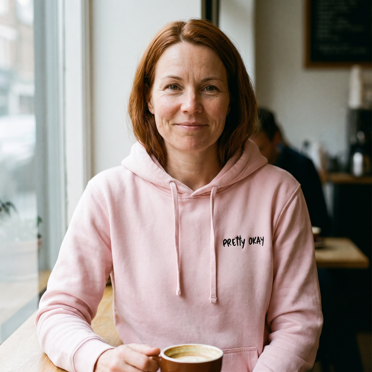 Pretty okay embroidered Finnish humour hoodie in light pink, worn by Sanna at a cafe window, Very Finnish Problems