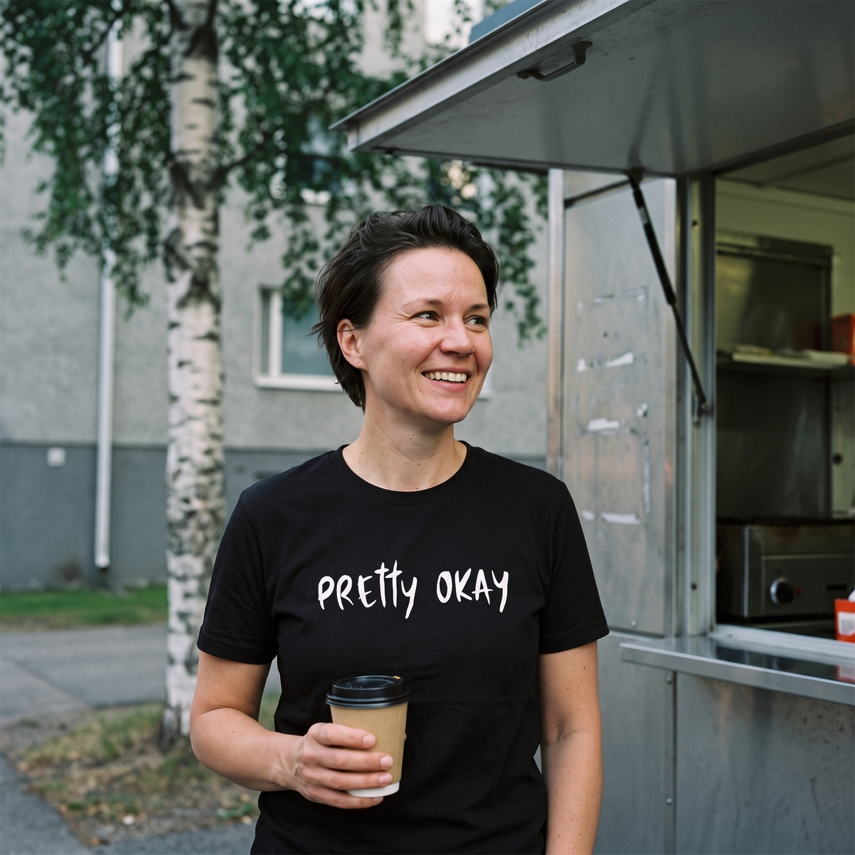 Pretty Okay t-shirt in black, worn by a woman with a coffee at a Finnish street grilli, Very Finnish Problems