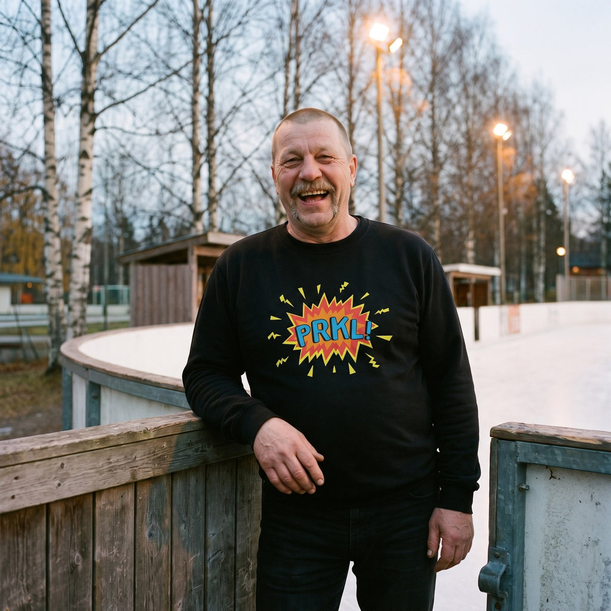 PRKL Bang sweatshirt in black, worn by Juha laughing by an outdoor ice rink, Very Finnish Problems