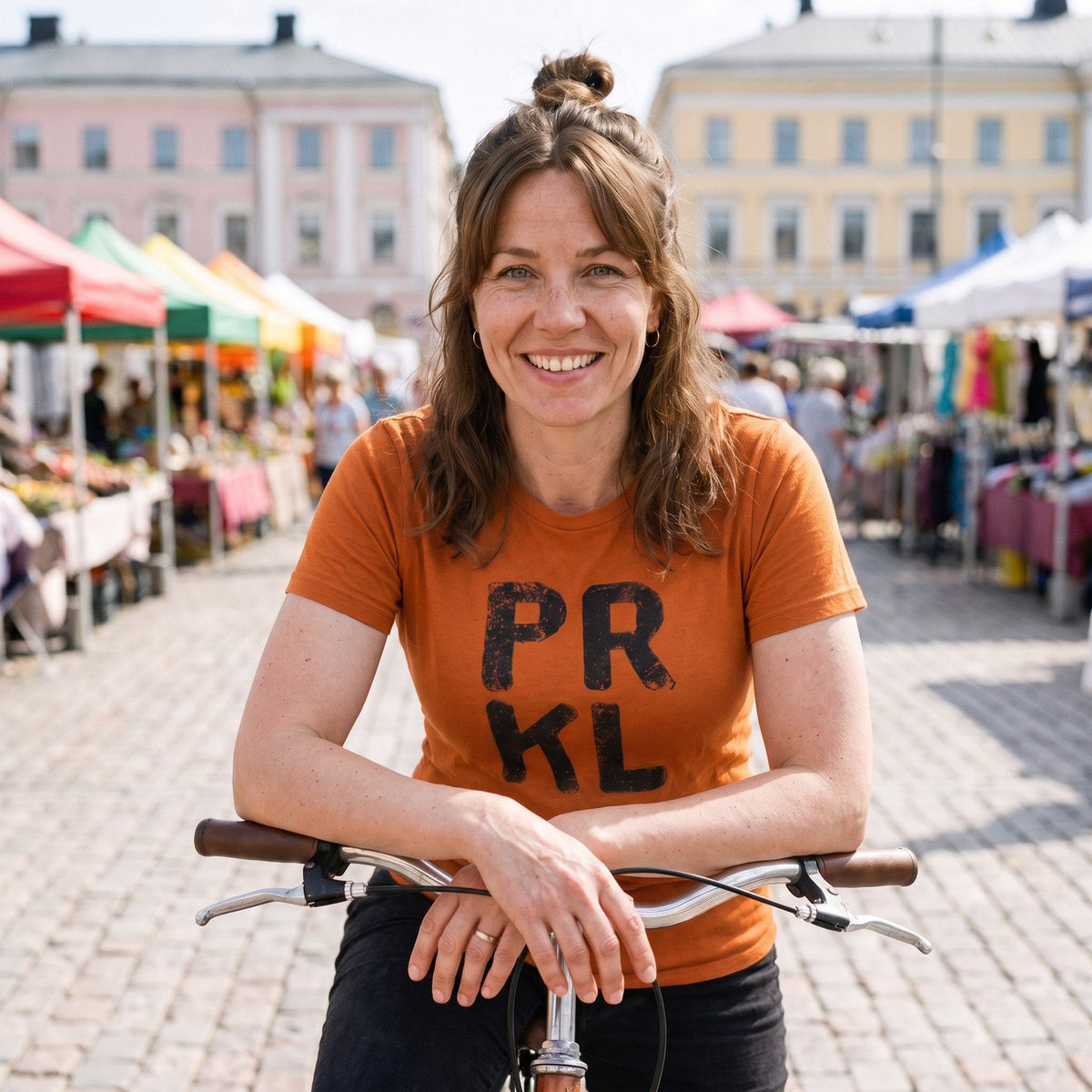 PRKL Finnish language t-shirt in burnt orange, worn by Suvi leaning on her bicycle at a Finnish market square, Very Finnish Problems