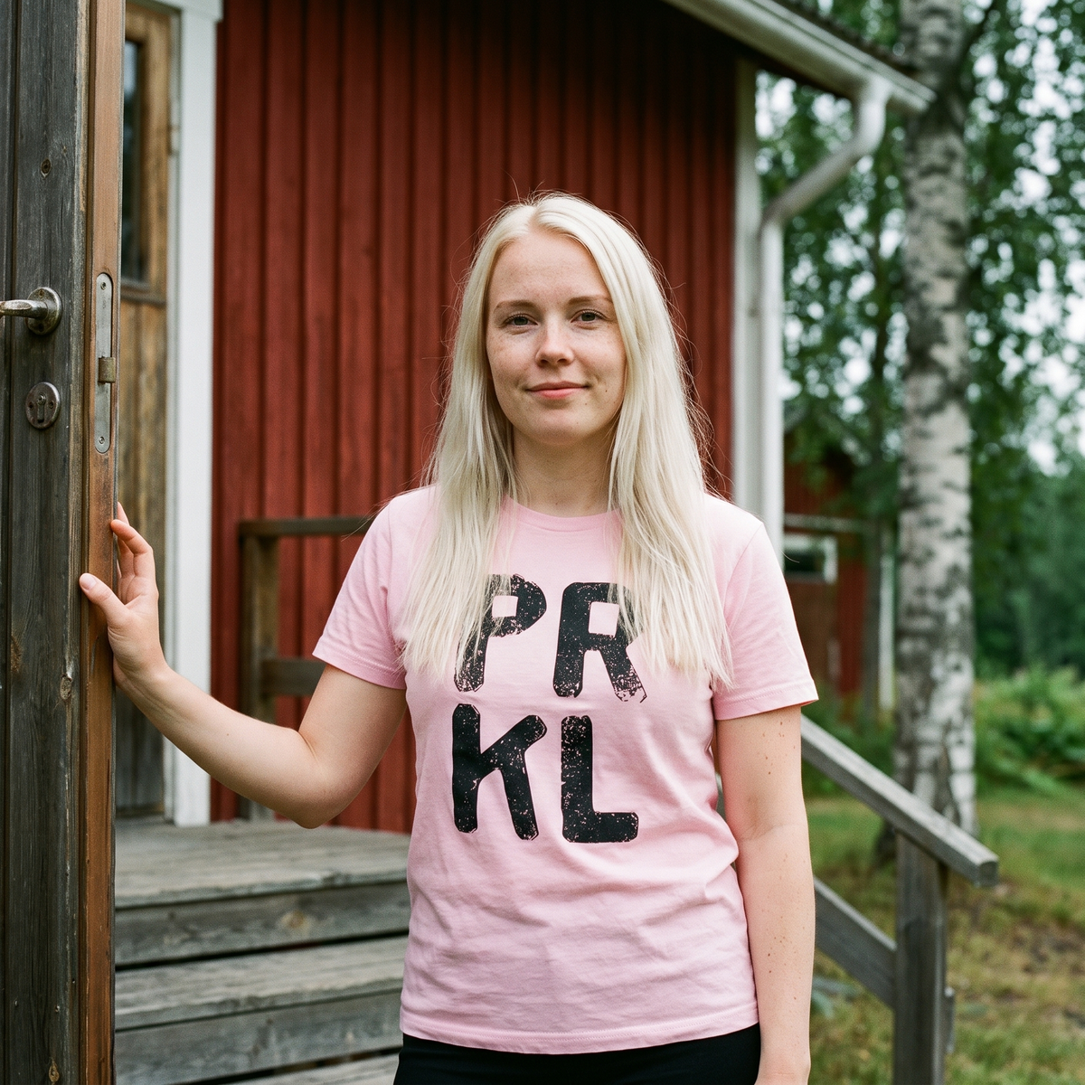 PRKL Finnish language t-shirt in pink, worn by a young woman on the steps of a red summer cottage, Very Finnish Problems