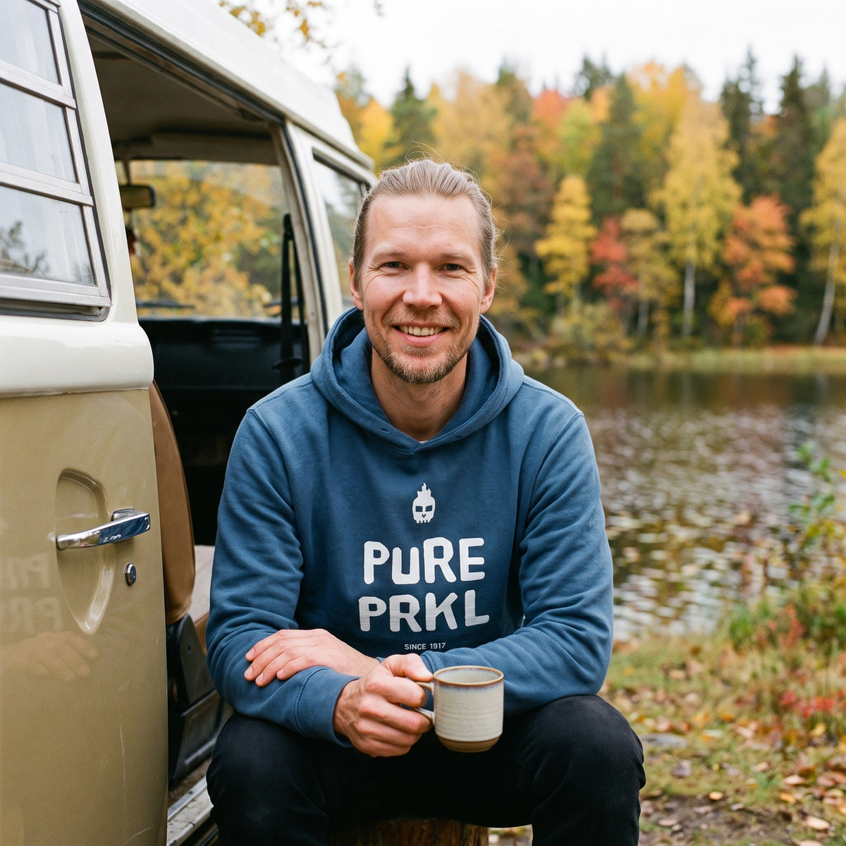 Pure PRKL hoodie in indigo blue, worn by Joonas in a camper van doorway by a lake, Very Finnish Problems