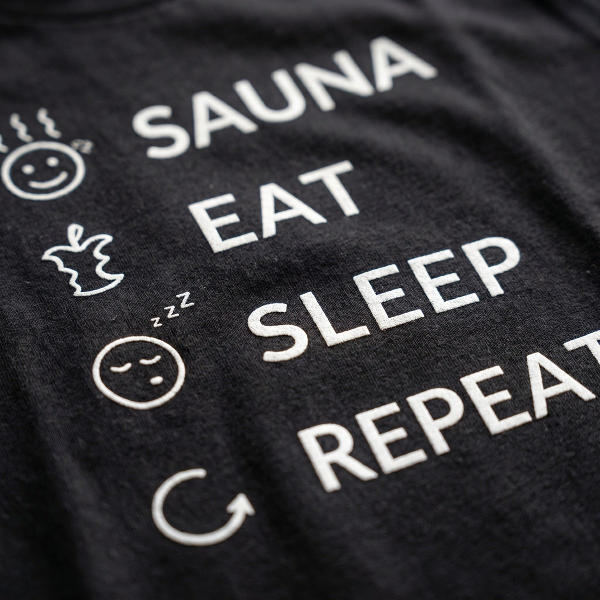 Close-up of the white Sauna Eat Sleep Repeat print on a black t-shirt, Very Finnish Problems