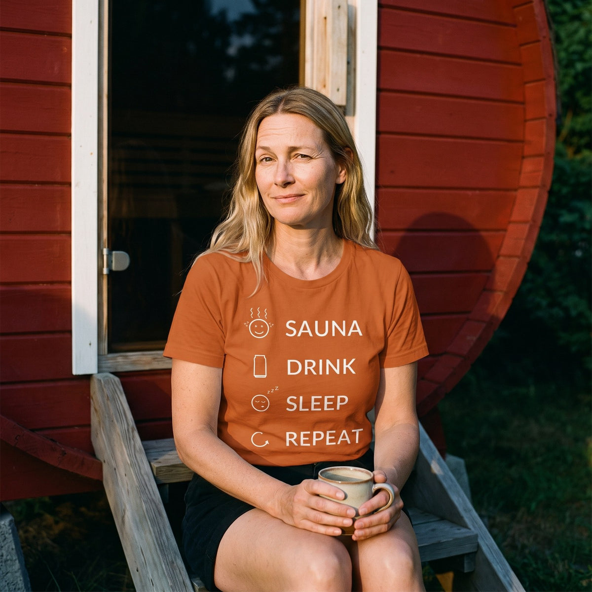 Sauna Drink Sleep Repeat t-shirt in autumn, worn by Elina with a coffee on the steps of a red sauna, Very Finnish Problems
