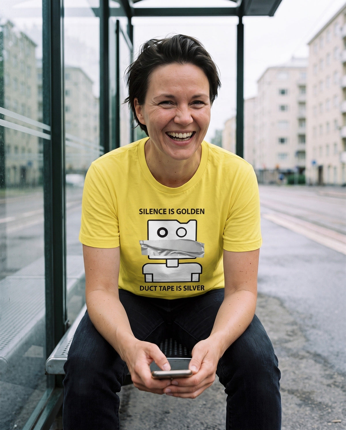 Silence is Golden Duct Tape is Silver t-shirt in yellow, worn by Katja laughing at her phone in a Helsinki tram shelter, Very Finnish Problems