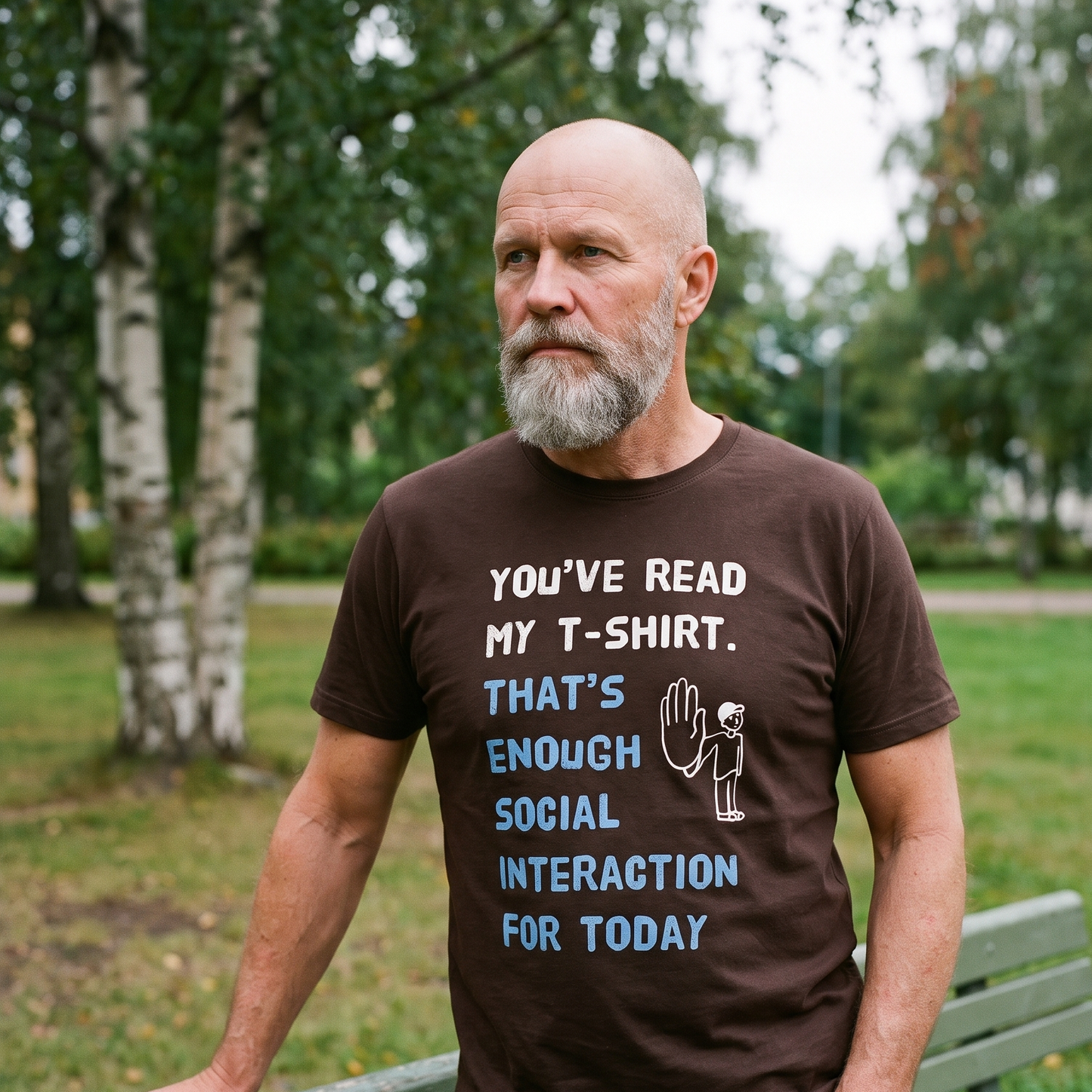 Social Interaction t-shirt in brown, worn by Mikko beside a park bench someone has just sat on, Very Finnish Problems