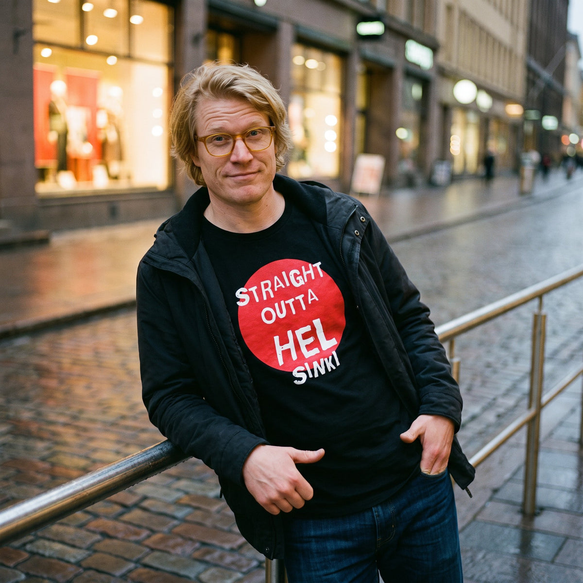 Straight Outta Hel t-shirt in black with a red circle, worn by Tuomas on a wet Helsinki street at dusk, Very Finnish Problems