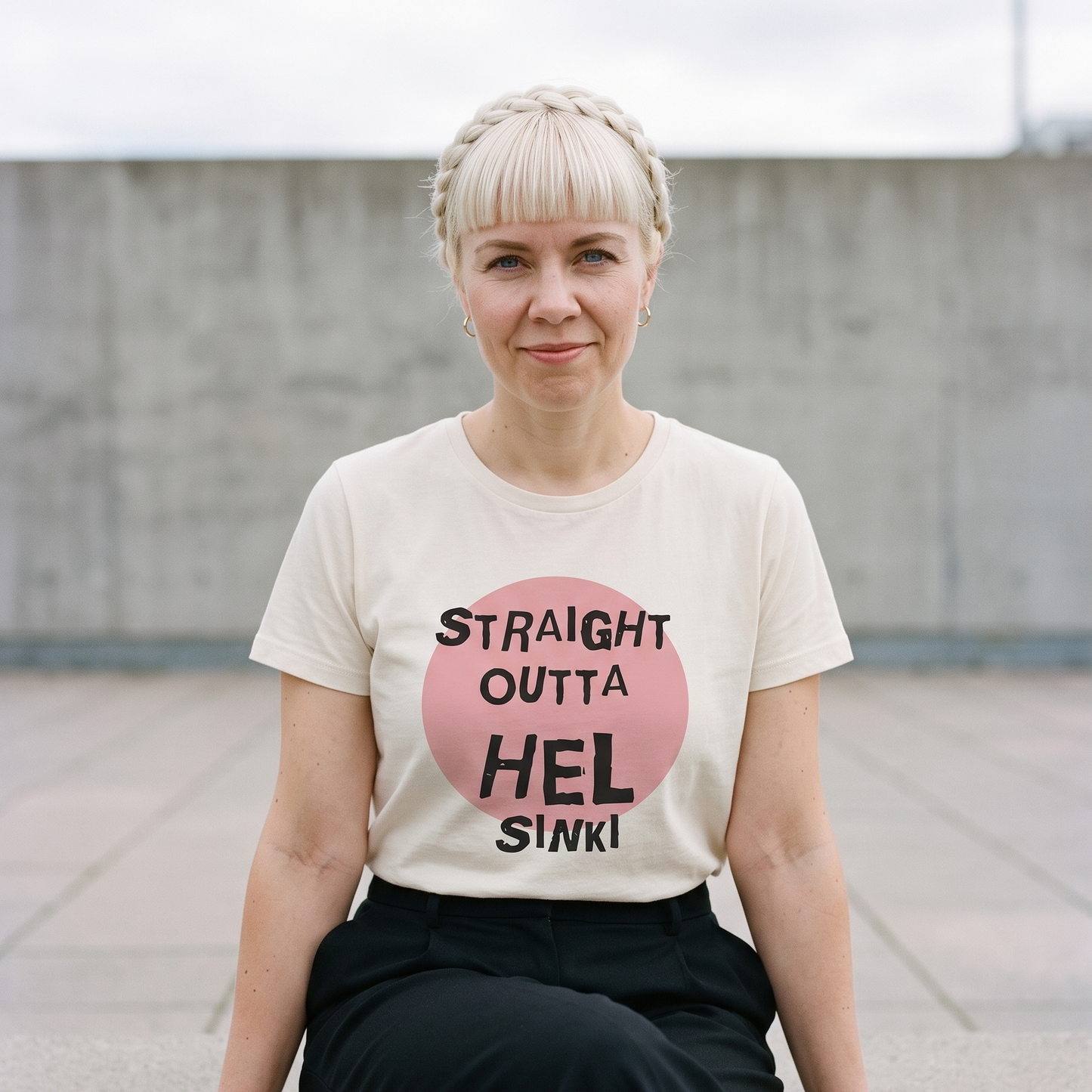 Straight Outta Hel t-shirt in soft cream, worn by Venla on a concrete ledge in a Finnish city plaza, Very Finnish Problems