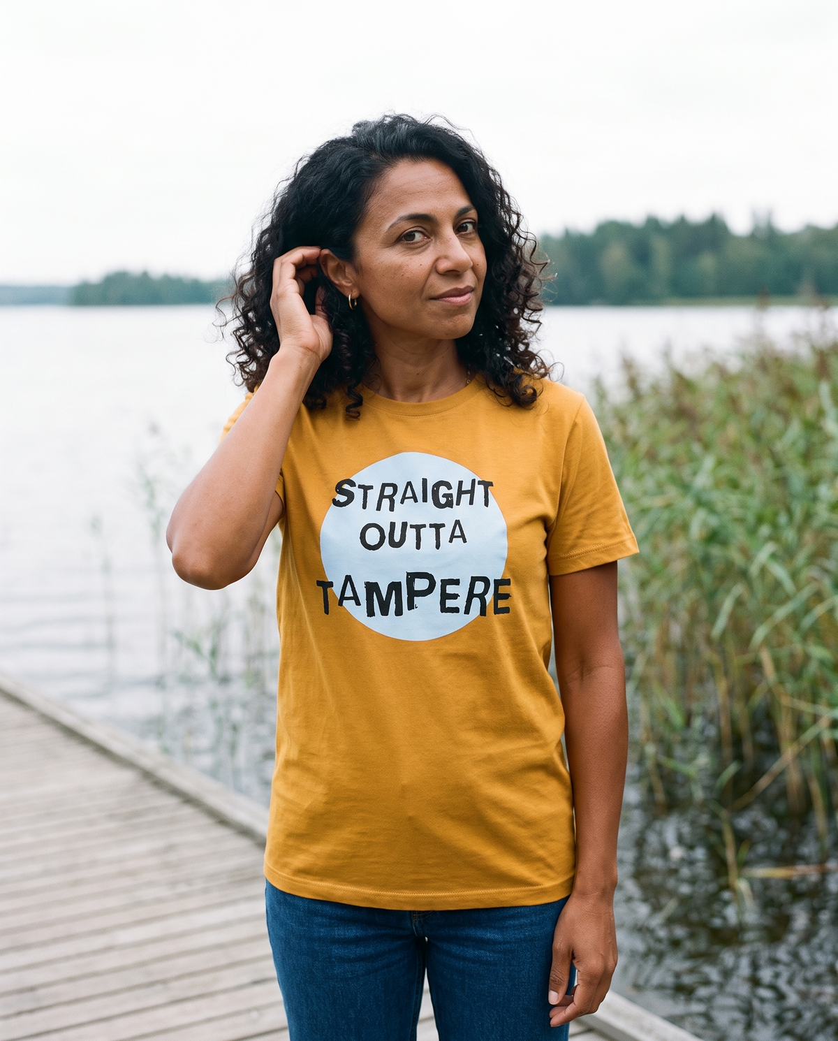 Straight Outta Tampere t-shirt in gold, worn by Nea on a lakeside boardwalk, Very Finnish Problems