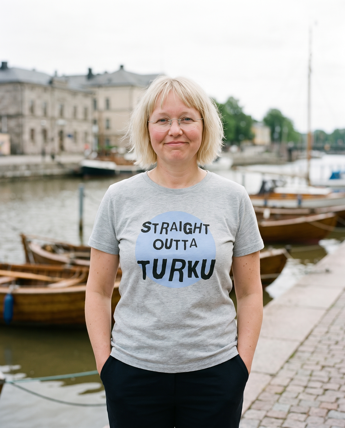 Straight Outta Turku t-shirt in silver, worn by Marika on a granite river quay, Very Finnish Problems