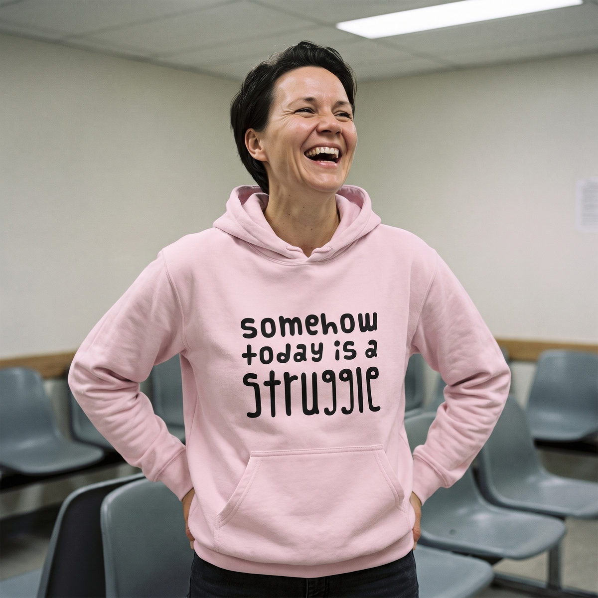 Somehow today is a struggle Finnish culture hoodie in light pink, worn by Katja laughing in a waiting room, Very Finnish Problems