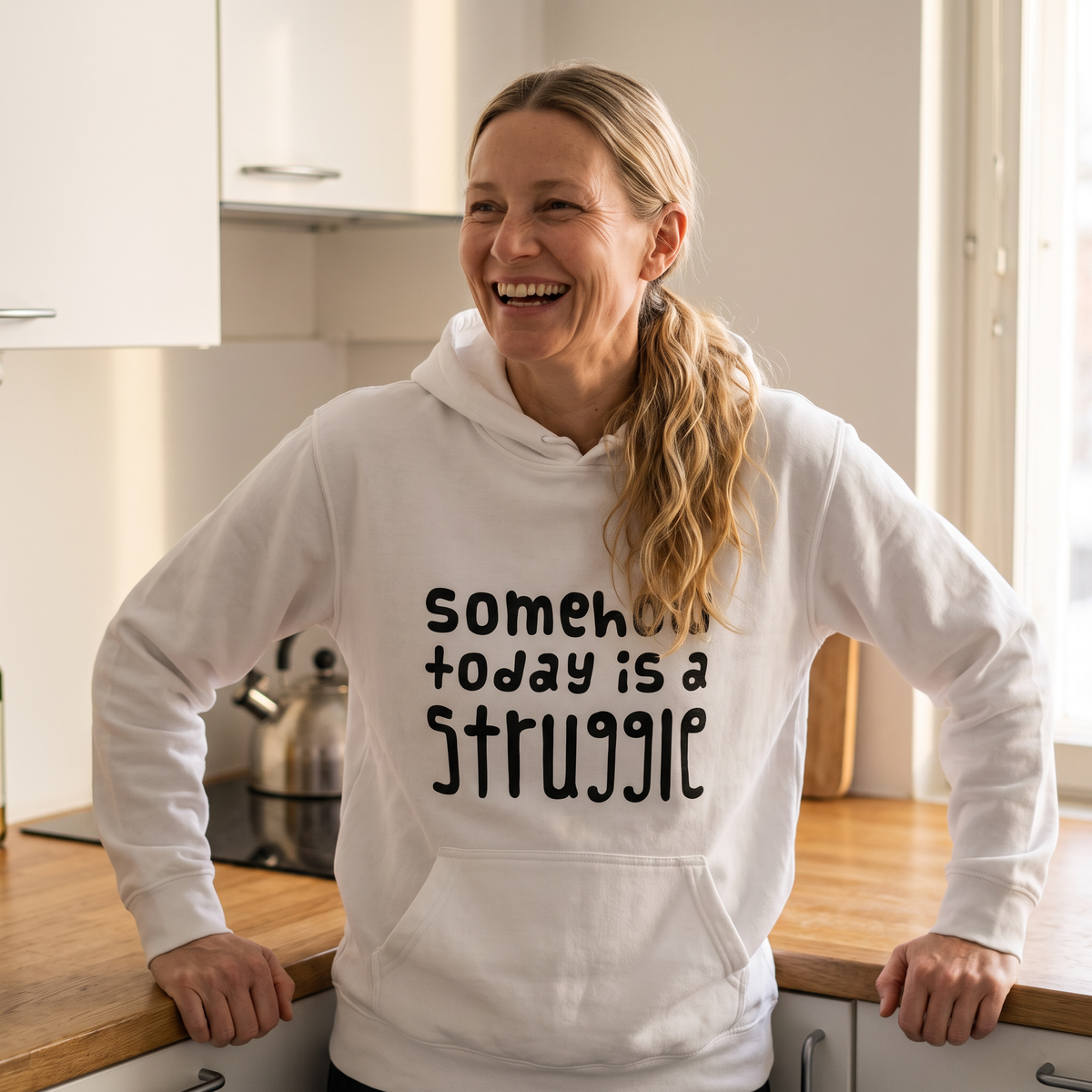 Somehow today is a struggle Finnish culture hoodie in white, worn by Elina laughing in a kitchen in the morning, Very Finnish Problems