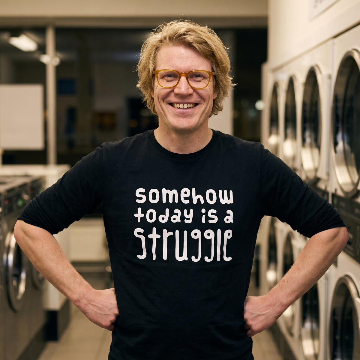 Somehow today is a struggle Finnish long sleeve tee in black, worn by Tuomas grinning in a launderette, Very Finnish Problems