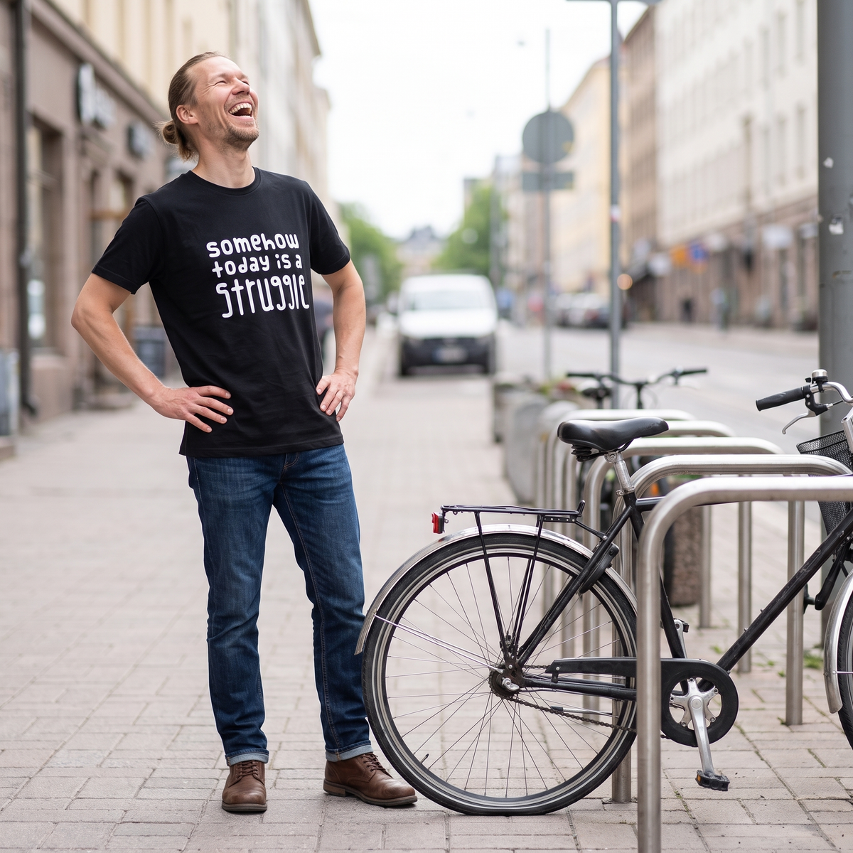Somehow today is a struggle Finnish organic cotton t-shirt in black, worn by Joonas laughing beside a bicycle with a flat tyre, Very Finnish Problems