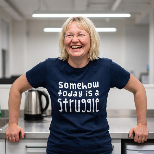 Somehow today is a struggle Finnish culture t-shirt in navy, worn by Marika laughing in an office kitchen, Very Finnish Problems