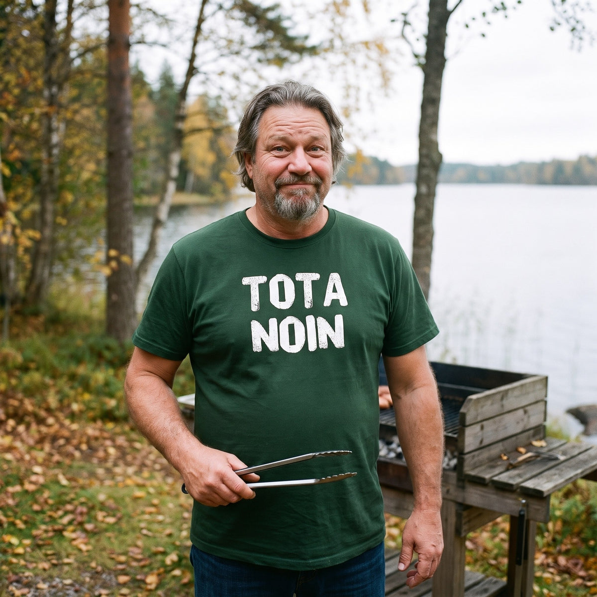 Tota noin Finnish language t-shirt in forest green, worn by a man hesitating with tongs at a lakeside grill, Very Finnish Problems
