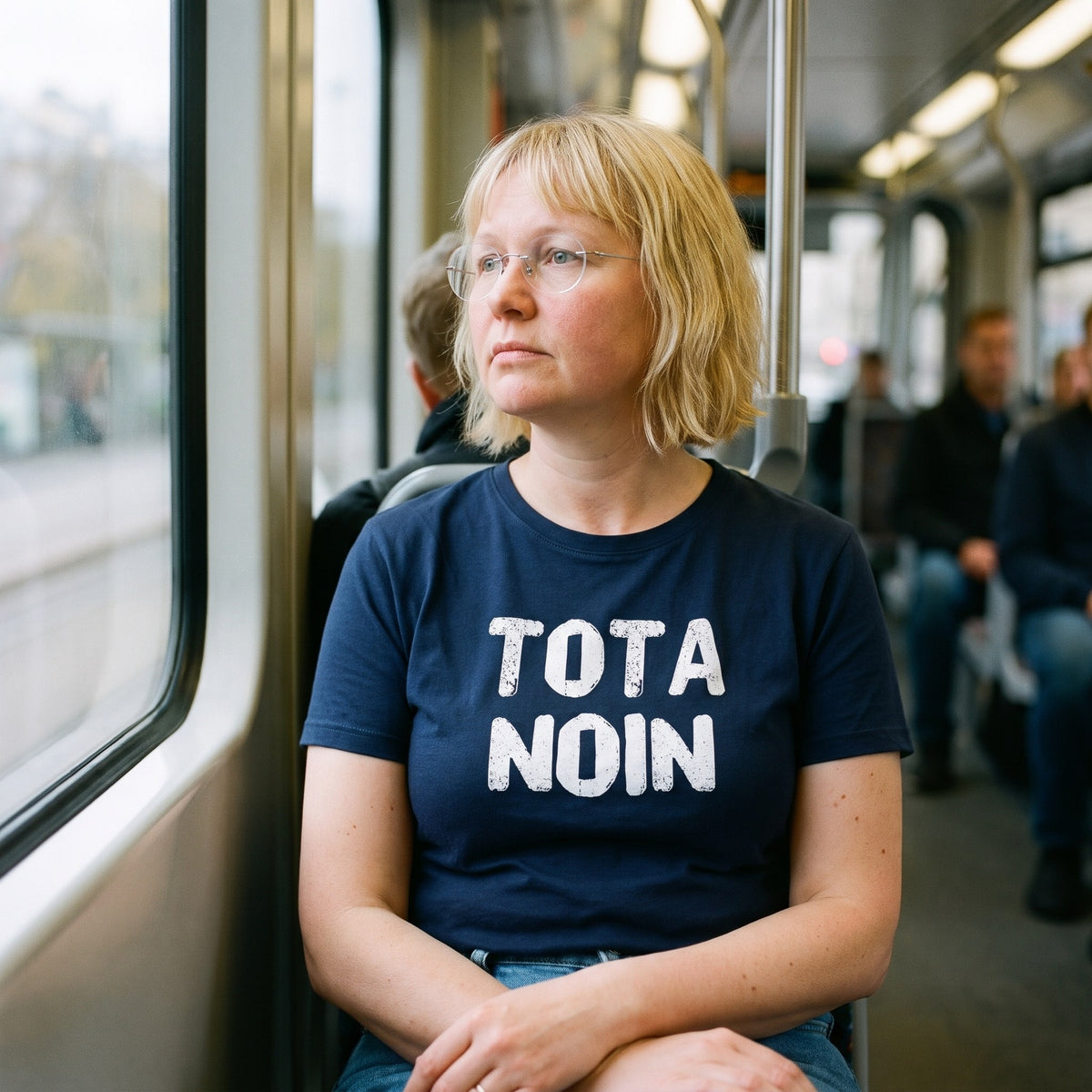 Tota noin Finnish language t-shirt in navy, worn by a woman lost in thought by a tram window, Very Finnish Problems