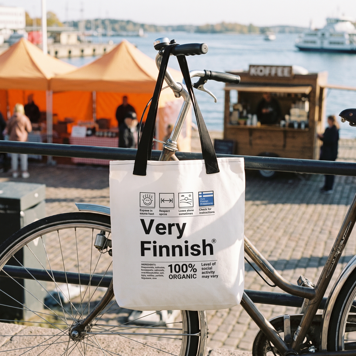 Very Finnish tote bag hanging from a bicycle at Helsinki market square, orange market tents and a coffee stall behind, Very Finnish Problems