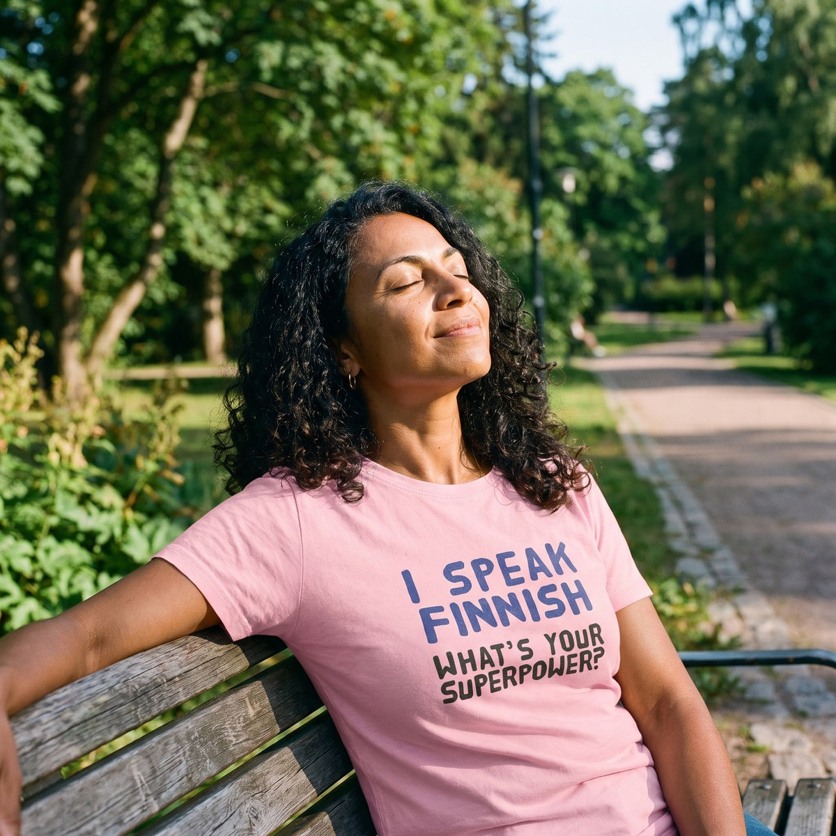 What's Your Superpower t-shirt in pink, worn by Nea in the sun on a Helsinki park bench, Very Finnish Problems