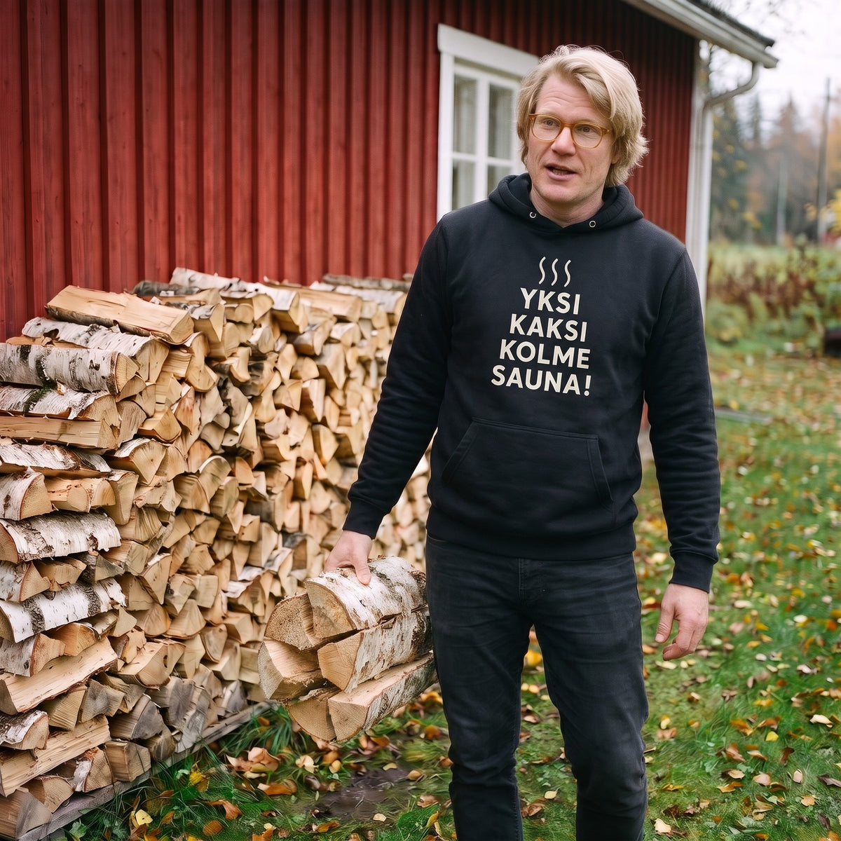 Yksi, kaksi, kolme, sauna! hoodie in black, worn by a man by the woodpile with an armful of birch logs, Very Finnish Problems