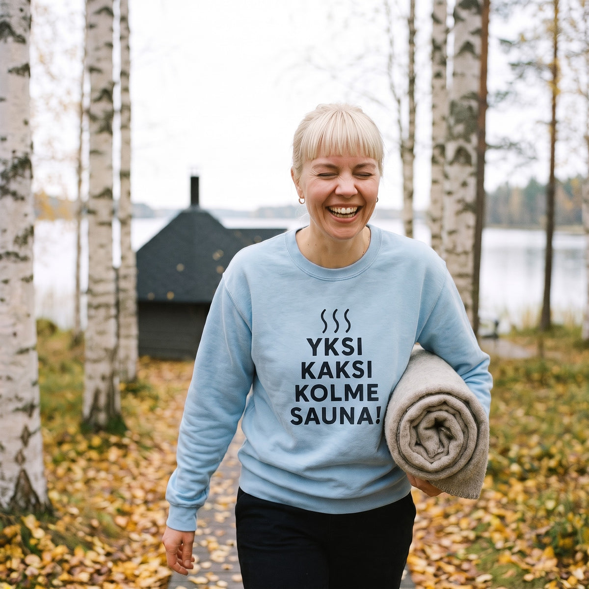 Yksi, kaksi, kolme, sauna! sweatshirt in light blue, worn by a laughing woman carrying a blanket down a birch path to a lakeside sauna, Very Finnish Problems