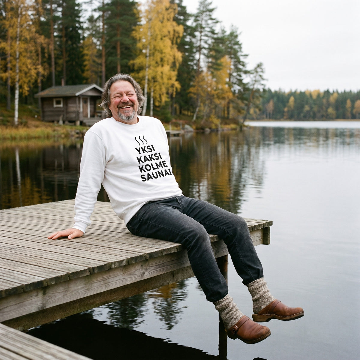 Yksi, kaksi, kolme, sauna! sweatshirt in white, worn by a grinning man sitting on a wooden lake jetty in autumn, Very Finnish Problems